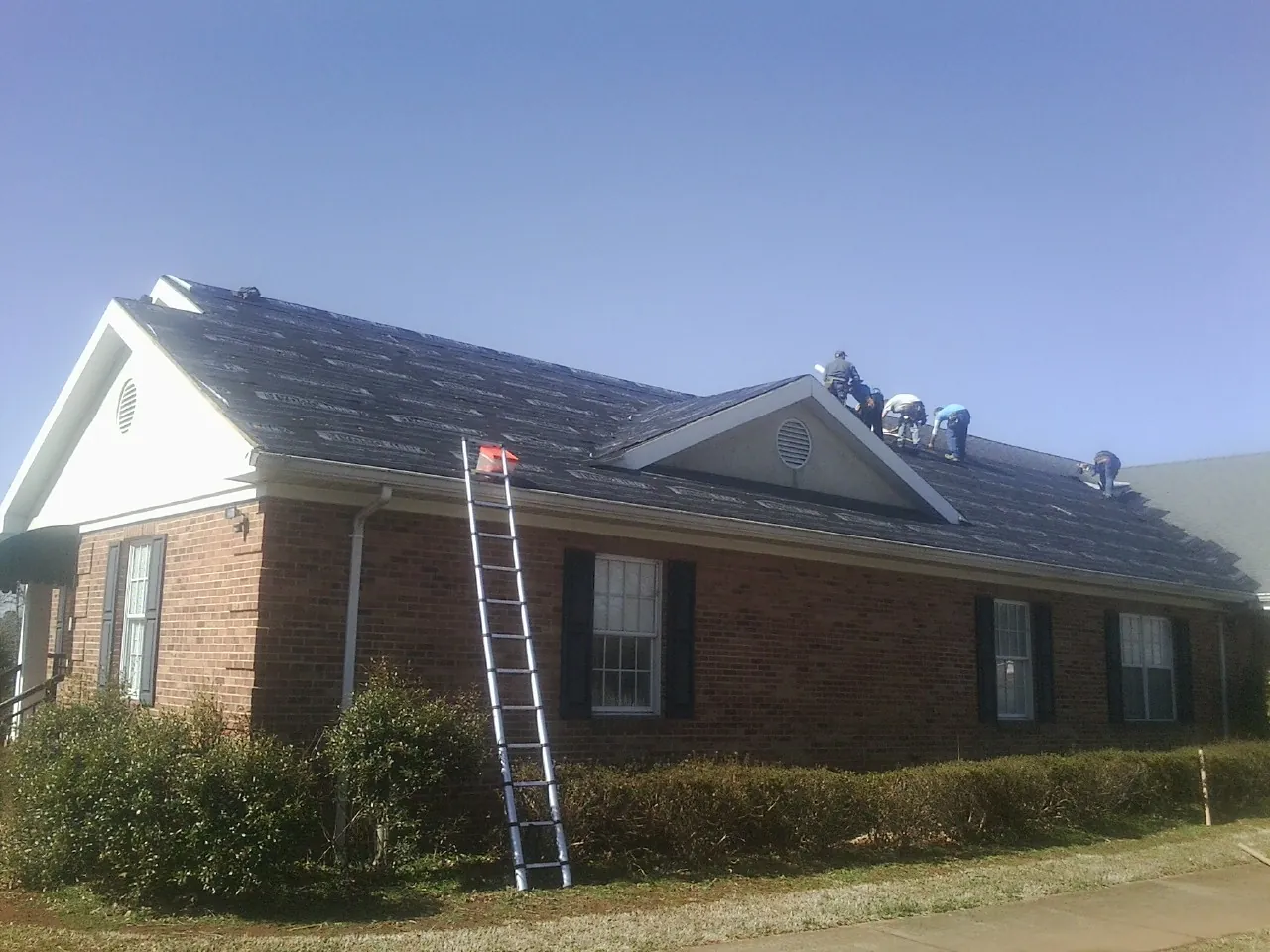 Crew tearing shingles off a brick ranch roof with a ladder set — Emergency Roof Repair work by our crew in Potsdam