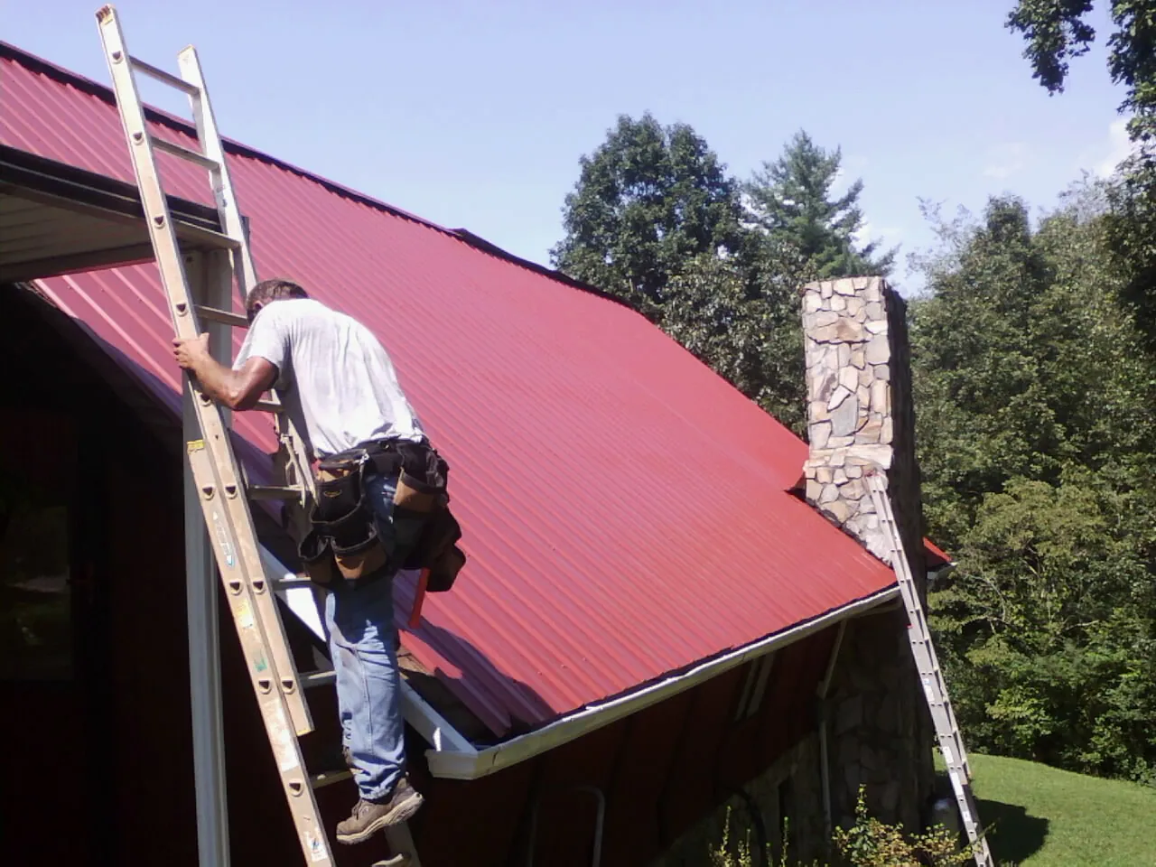 Roofer on a ladder beside a red metal roof and stone chimney — Soffit & Fascia Repair by Roofing Brothers in Potsdam